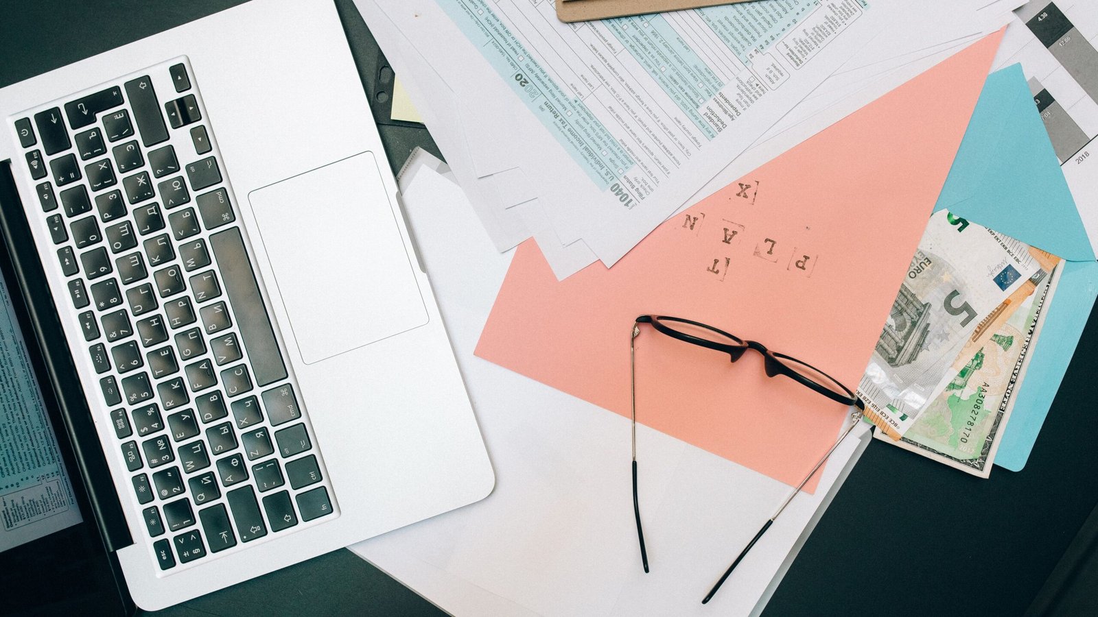 Laptop, paperwork, glasses and currency arranged at a desk for tracking client invoice payments
