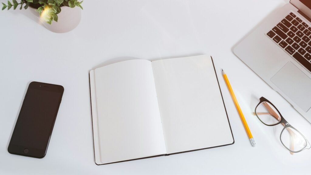 Open blank notebook beside a laptop, phone, pencil and glasses.