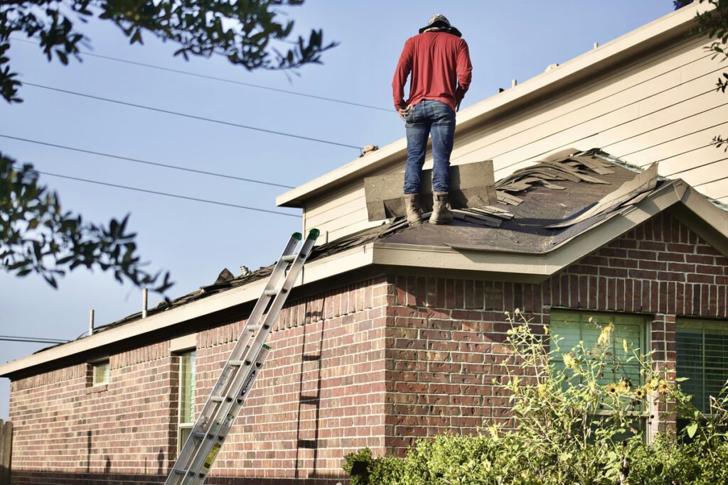 A worker stands on a roof beside loose shingles and a ladder.