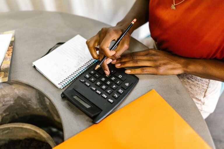 A person holds a pencil while pressing a calculator beside a spiral notebook on a gray table.