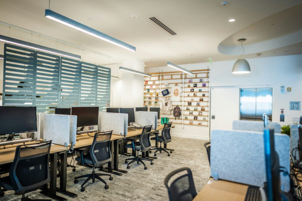 An empty office has desks, dark computer monitors, chairs and light gray desk dividers.