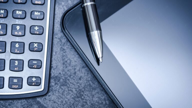 A silver pen rests on a tablet beside a calculator on a textured gray surface.