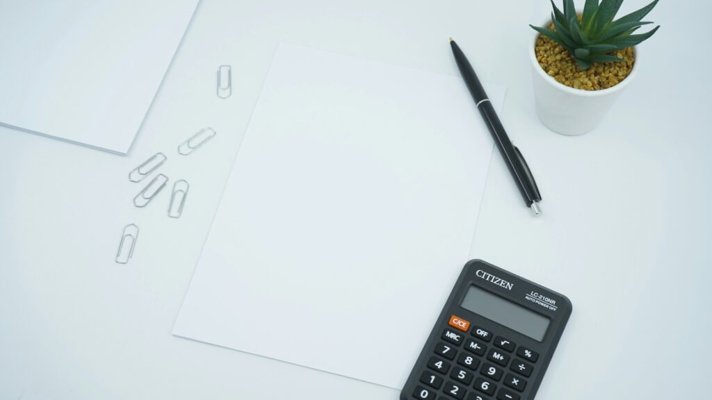 Blank paper, a pen and a calculator sit on a white desk beside paper clips and a small plant.