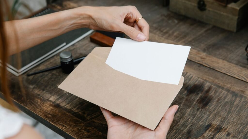 Hands taking a blank white card from a brown envelope over a wooden desk.