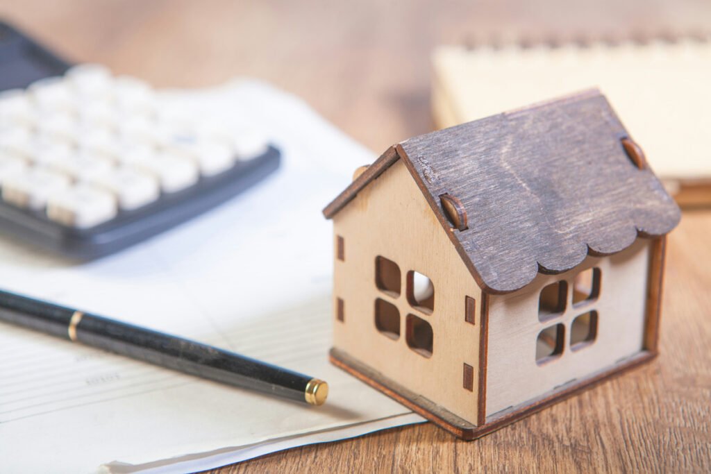A wooden house model beside a pen, paper and calculator.