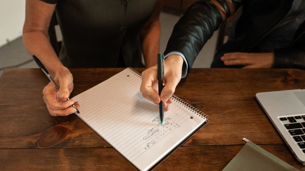 Two people hold pens above an open notebook on a wooden table beside a laptop.