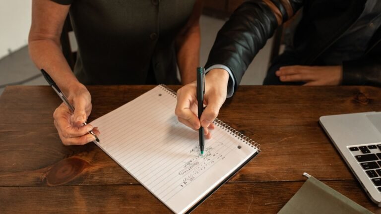Two people hold pens above an open notebook on a wooden table beside a laptop.