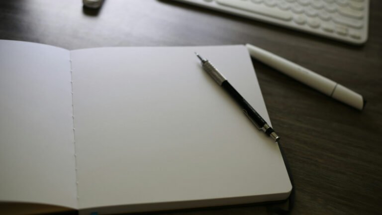An open blank notebook, pencil and pen beside a keyboard on a wooden desk.