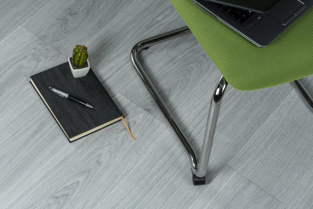 A pen and potted cactus rest on a closed notebook on a wood-patterned floor beside a green chair holding a laptop.