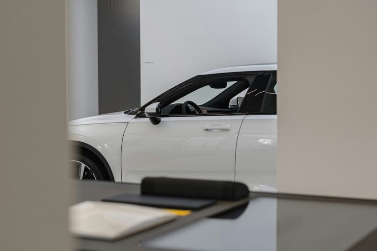 A white car is visible through an opening behind a dark desk in a bright showroom.