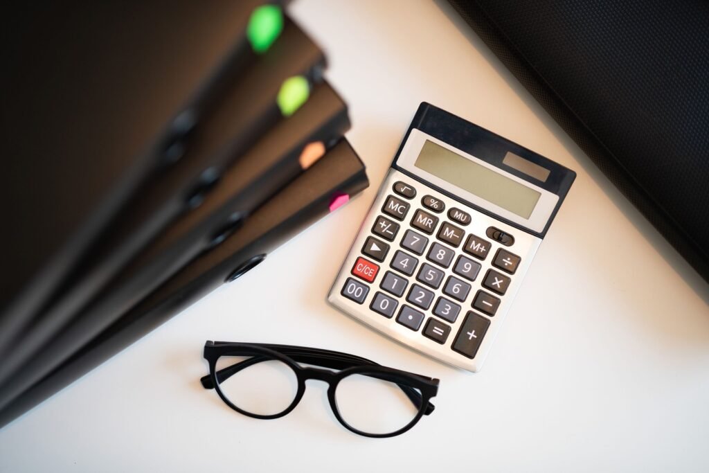 A calculator with a blank display and black-framed glasses sit beside closed folders with colored tabs.