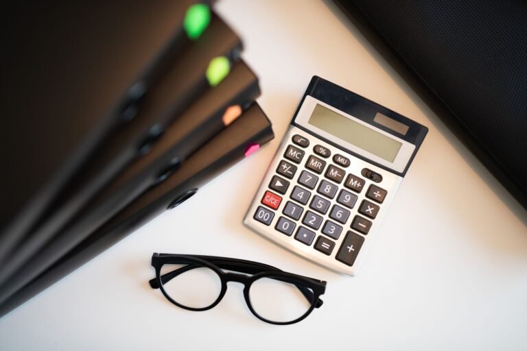 A calculator with a blank display and black-framed glasses sit beside closed folders with colored tabs.