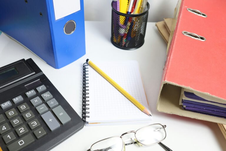 A calculator, blank squared notebook and yellow pencil sit between colorful binders on a desk.