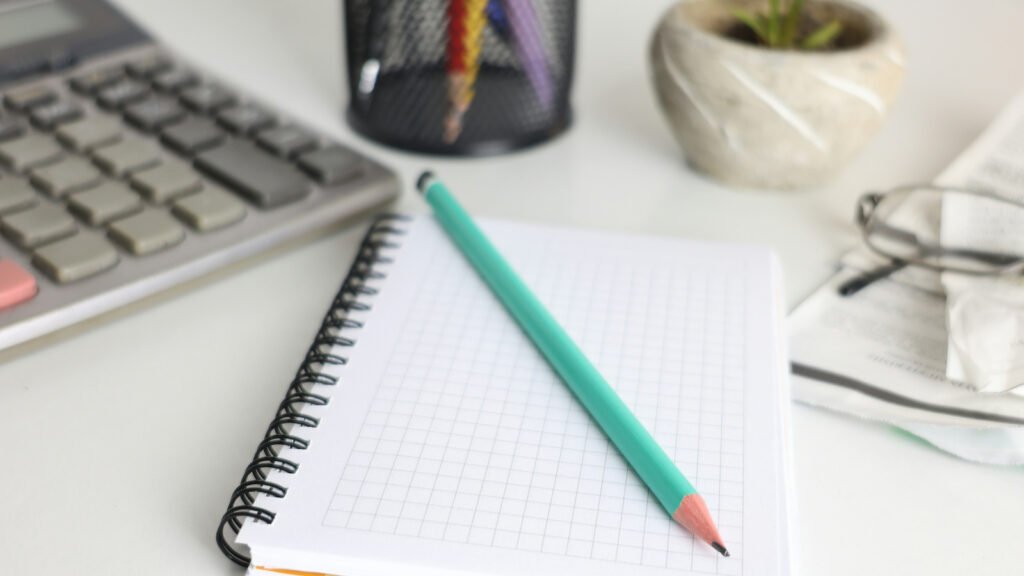 A teal pencil lies diagonally across a blank squared notebook beside a calculator and glasses.