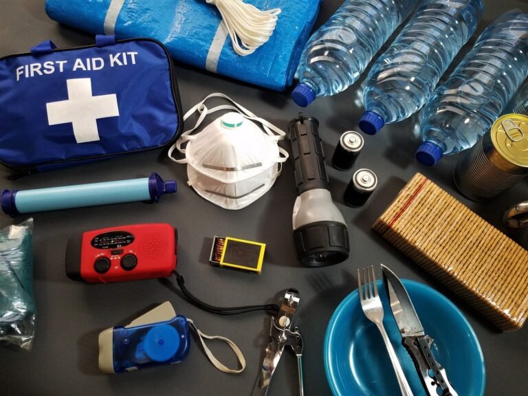 Bottled water, a first-aid kit, flashlight, batteries, portable radio and food arranged on a dark surface.