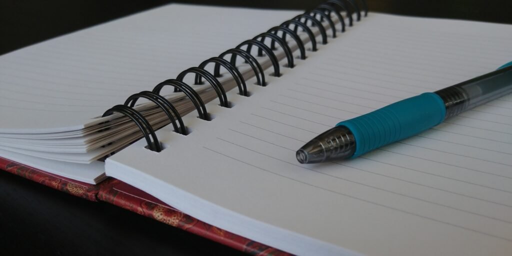 A blue pen resting on the blank lined pages of an open spiral notebook.