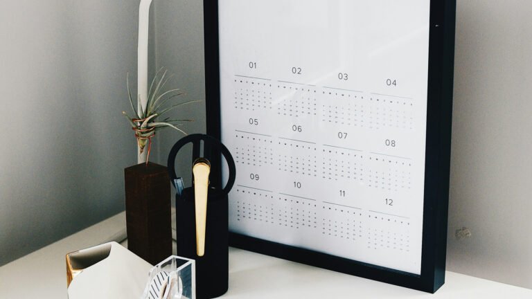 A framed annual calendar sits on a desk beside a small plant and stationery.