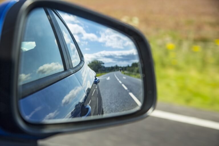 A car side mirror reflecting the blue car, a road and clouds.