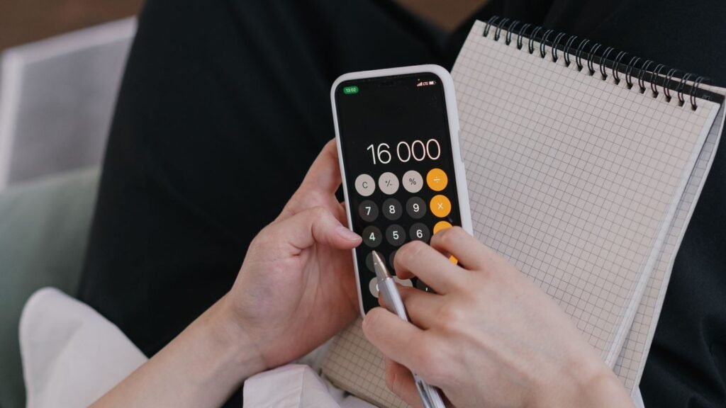 Hands using a smartphone calculator beside a blank grid notebook.