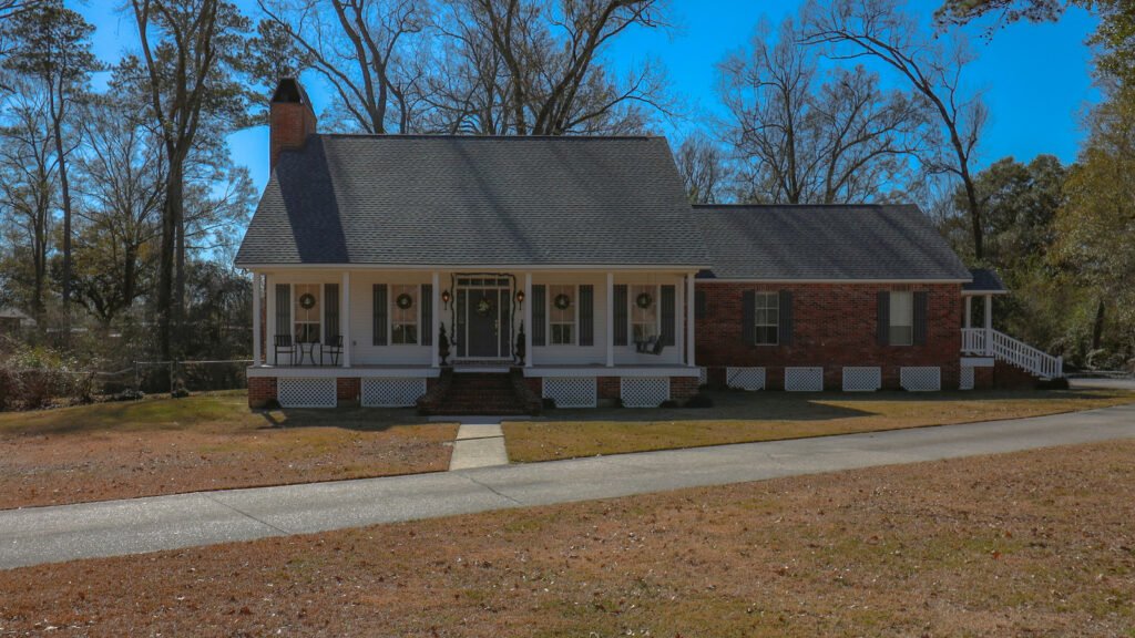 A brick and white-sided house has a front porch, lawn and trees.