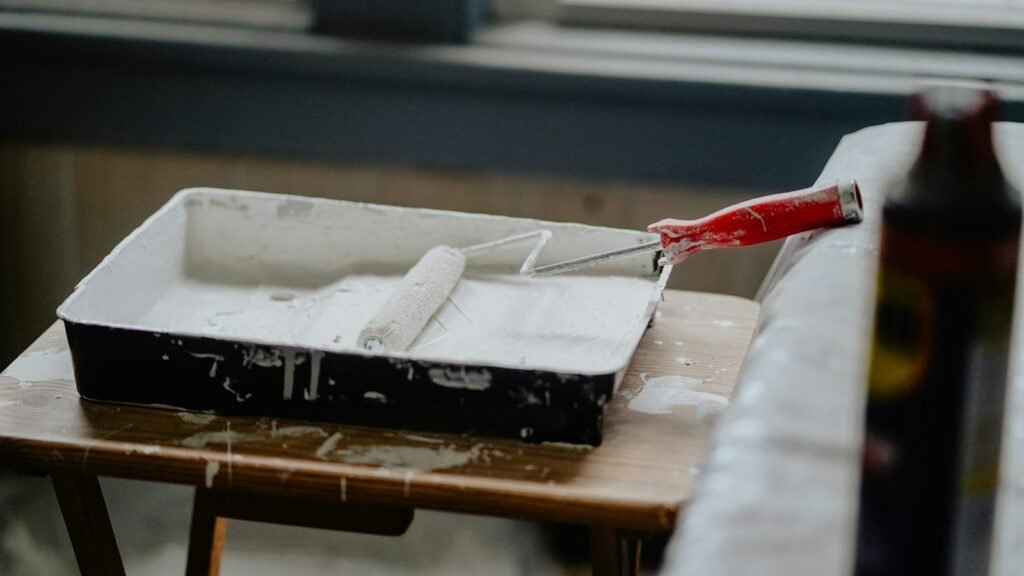 A red-handled paint roller rests in a tray of white paint.