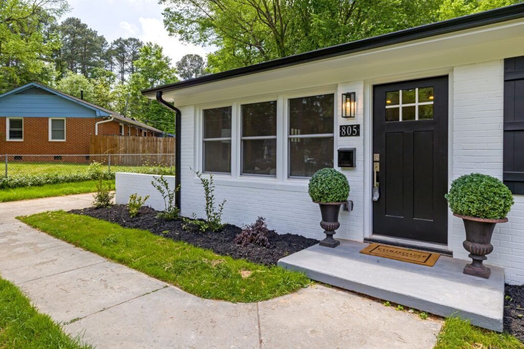 A white brick home has a black front door, potted shrubs and a concrete walkway beside a lawn.