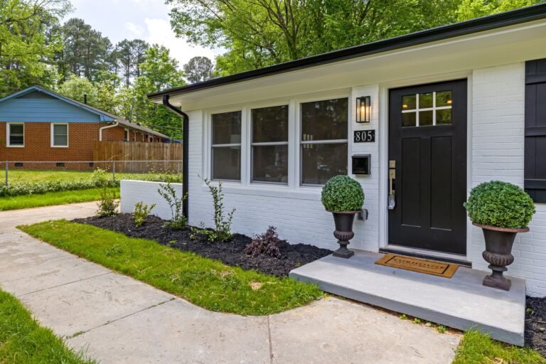 A white brick home has a black front door, potted shrubs and a concrete walkway beside a lawn.