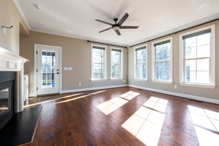 Sunlight crosses the hardwood floor of an empty living room with large windows, a fireplace and a ceiling fan.