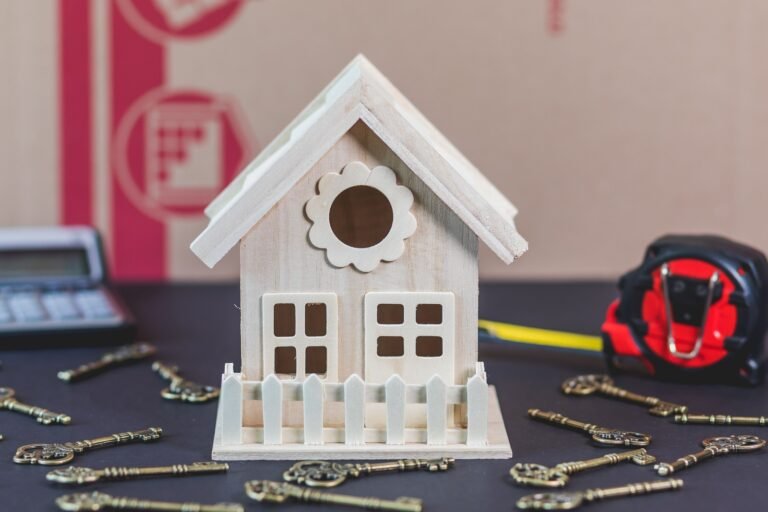 A miniature wooden house and decorative keys on a dark table, with a calculator and tape measure nearby.