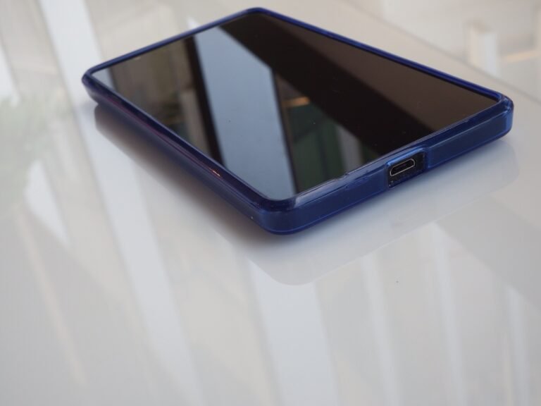 A smartphone in a blue case rests on a light reflective surface with its screen blank.