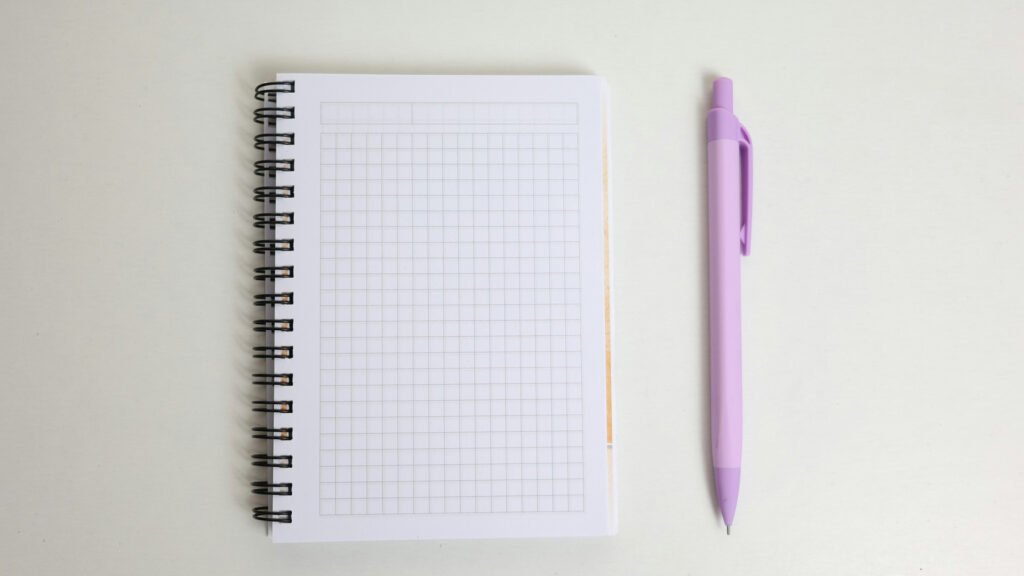 A blank grid-paper spiral notebook and purple pen on a pale desk.