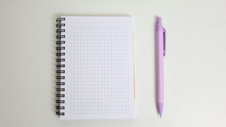A blank grid-paper spiral notebook and purple pen on a pale desk.