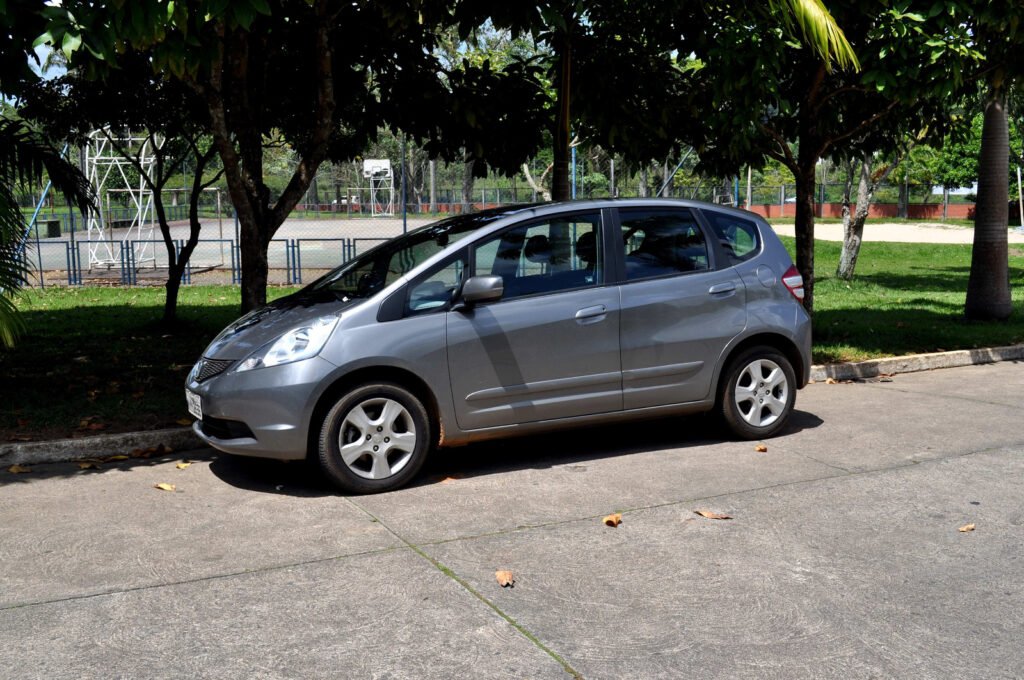 A silver hatchback is parked along a curb beside trees and a fenced sports court.