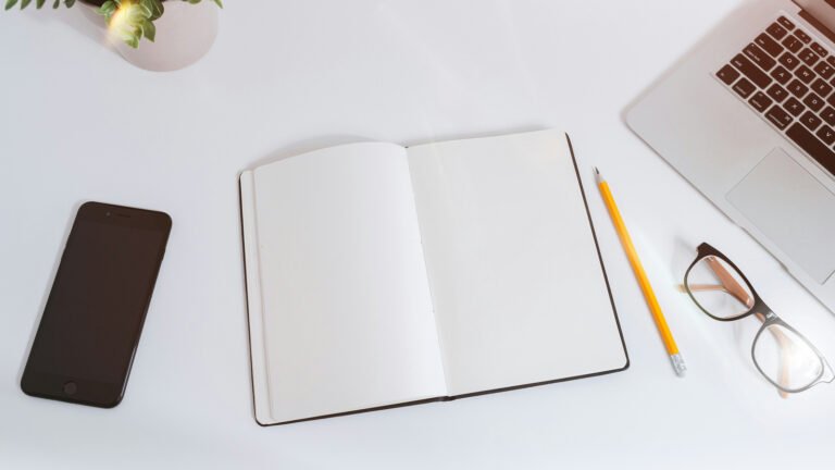An open blank notebook, pencil, phone, glasses and part of a laptop sit on a white desk.