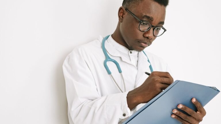 A man in a white medical coat and stethoscope writing on a clipboard.