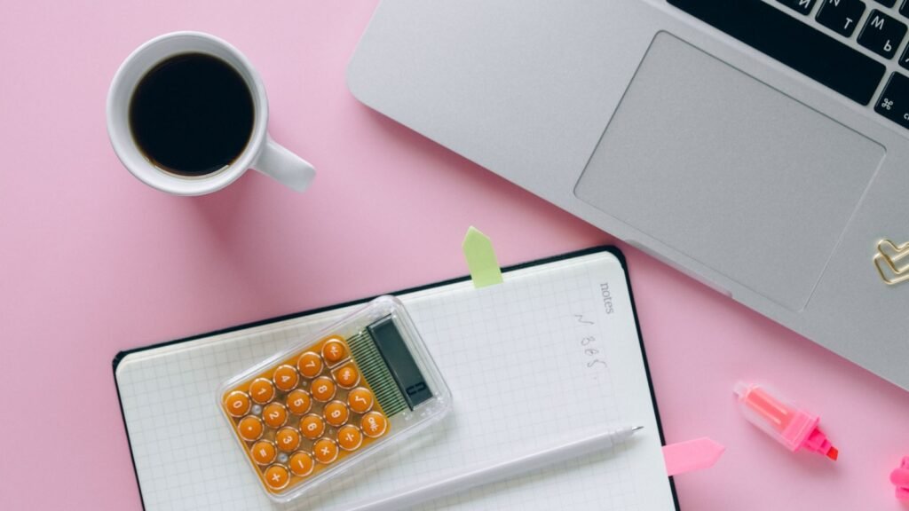 Calculator on an open notebook beside a laptop and coffee on a pink desk.