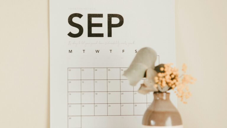 A paper wall calendar beside a vase of dried flowers.