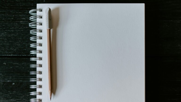 A wooden pencil rests beside the binding of a blank spiral notebook on a dark table.