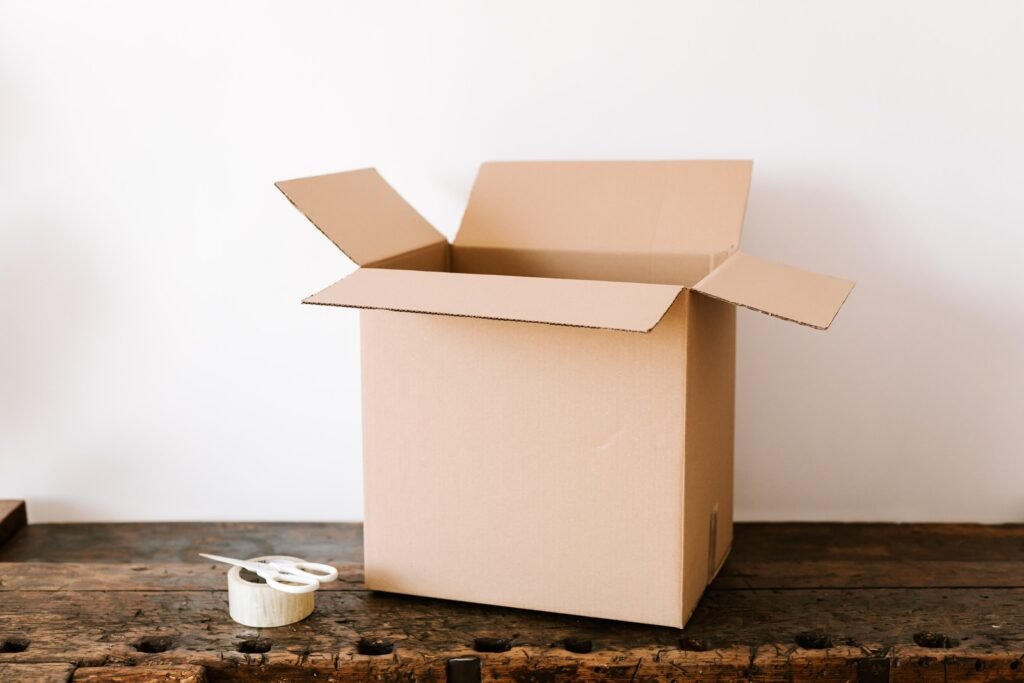 Open cardboard box beside clear packing tape and white scissors on a wooden table.