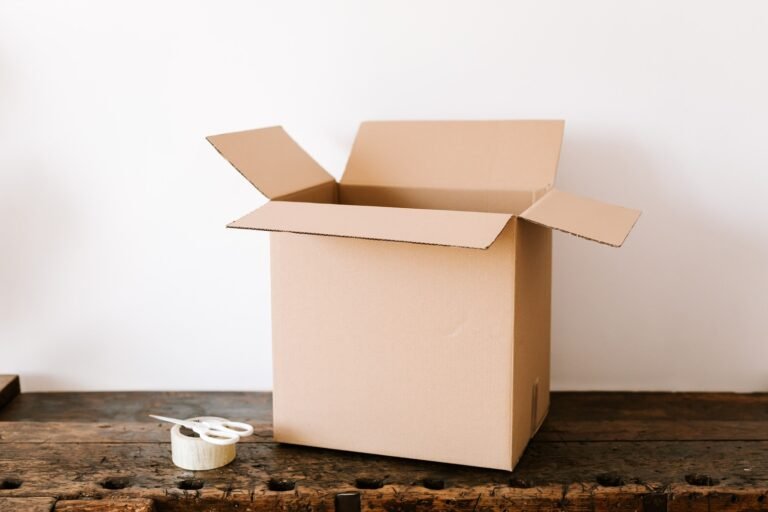 Open cardboard box beside clear packing tape and white scissors on a wooden table.