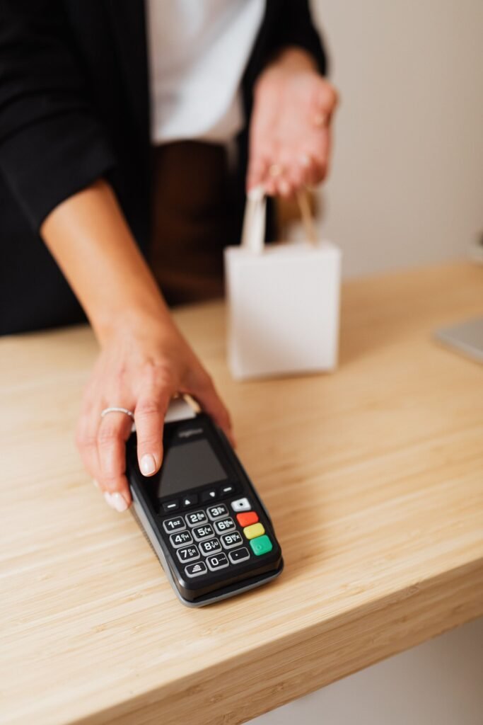 A hand holds a payment terminal beside a plain shopping bag on a wooden counter.