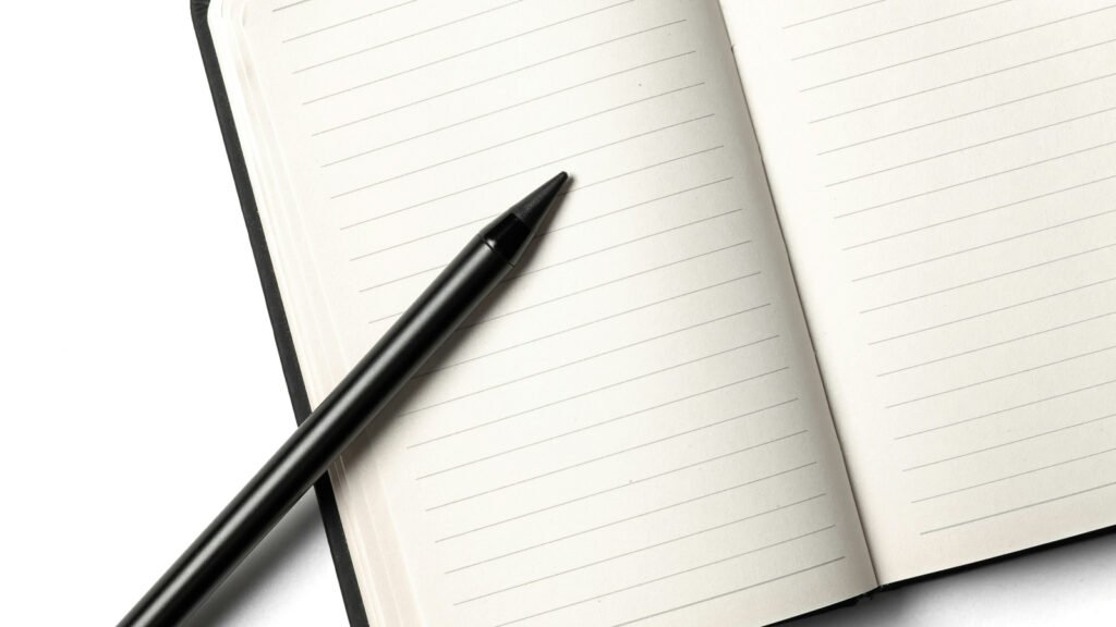 A black pen lies across an open blank lined notebook on a white surface.