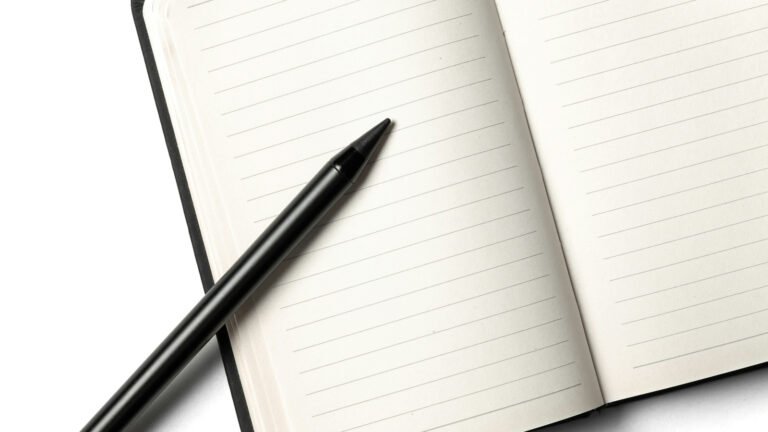 A black pen lies across an open blank lined notebook on a white surface.