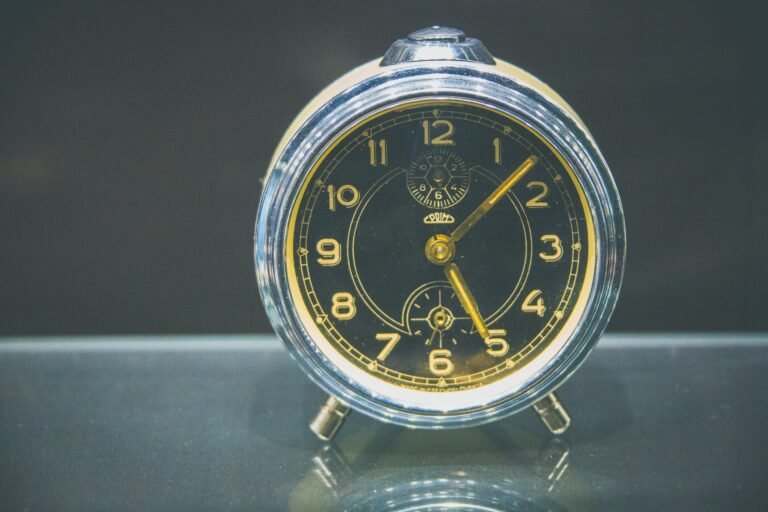 A vintage alarm clock with yellow numerals on a dark dial, standing on a reflective surface.