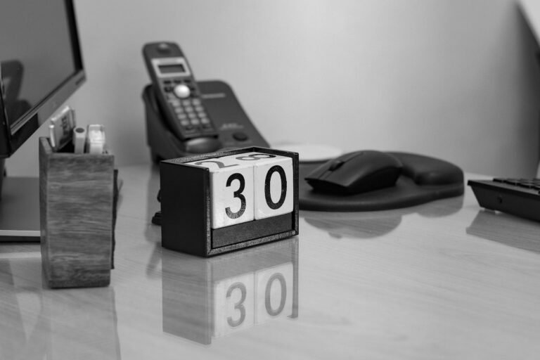 A black-and-white photograph of a desktop block calendar displaying 30, with a telephone and computer equipment behind it.