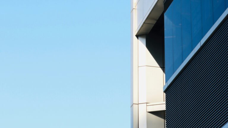 Modern building facade with blue windows, pale cladding and dark horizontal louvers against a clear sky.