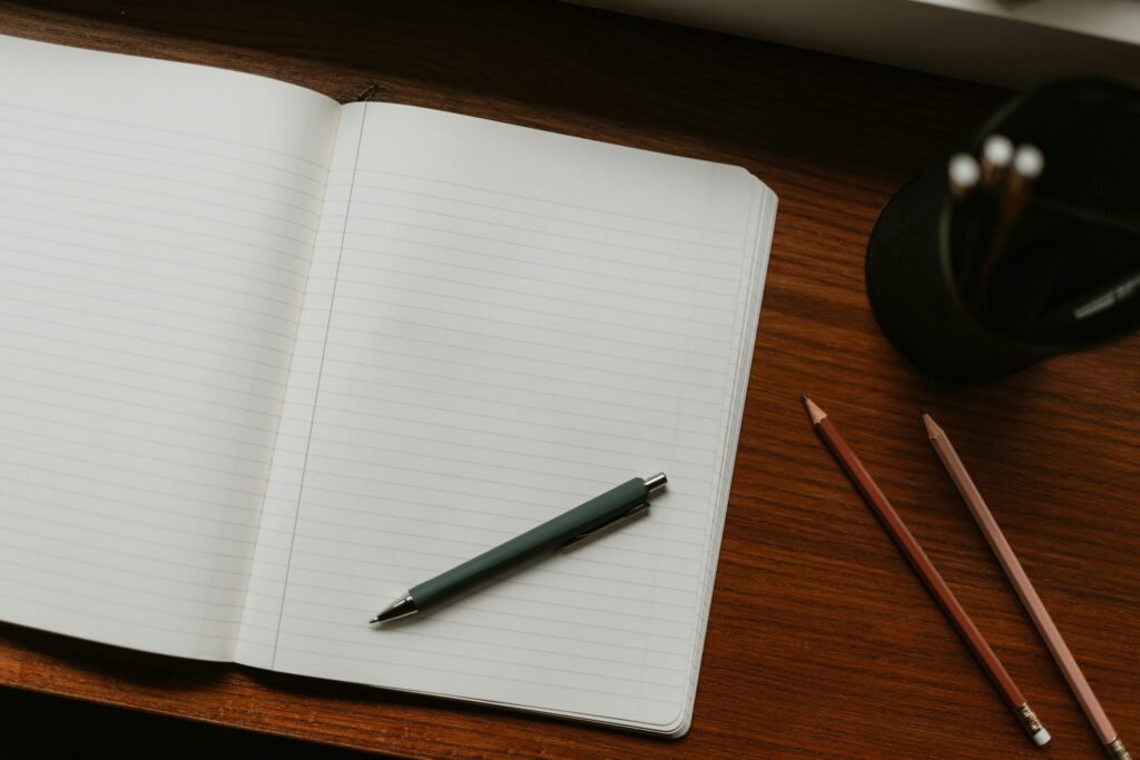 Blank lined notebook with a pen, pencils and a pencil cup on a wooden desk.