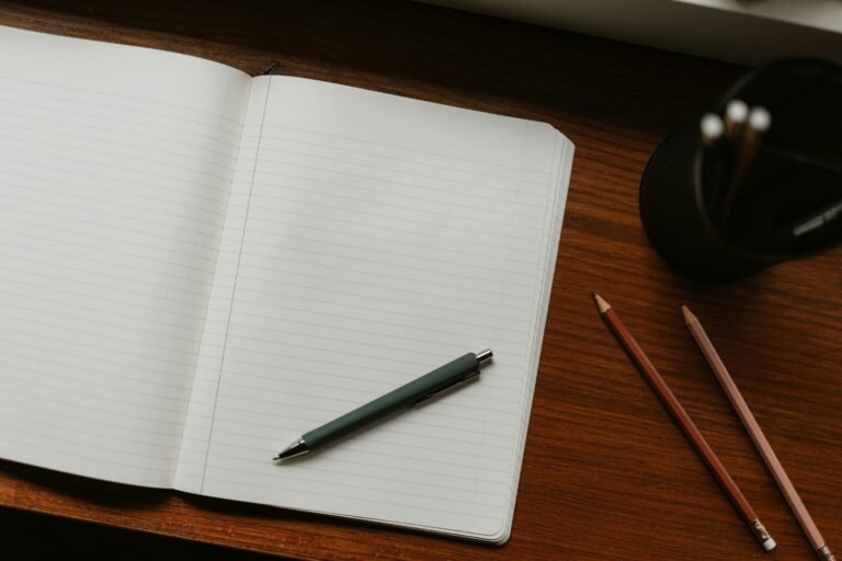 Blank lined notebook with a pen, pencils and a pencil cup on a wooden desk.