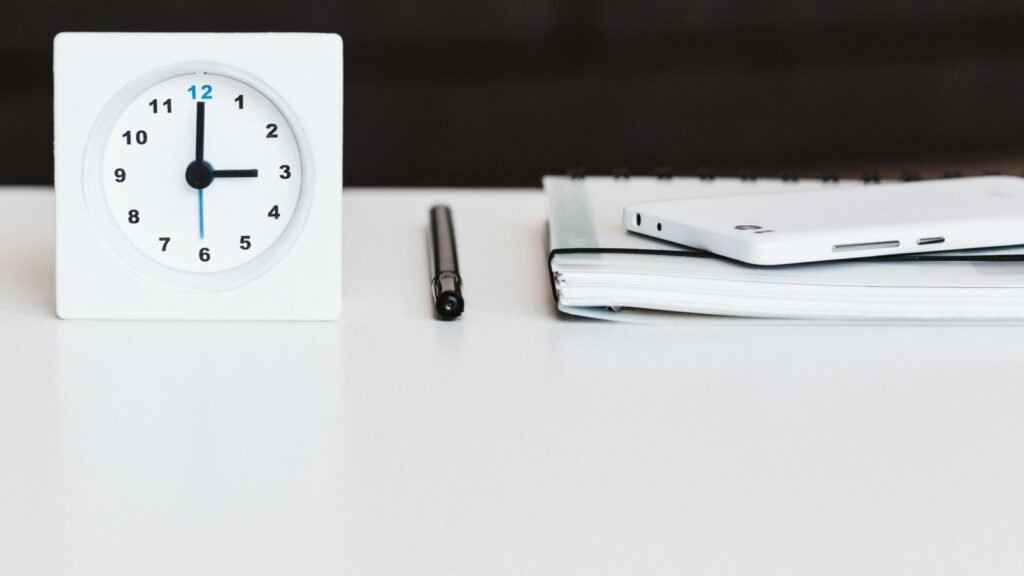 A white desk clock beside a pen, notebook and phone.