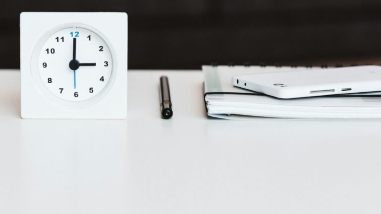 A white desk clock beside a pen, notebook and phone.